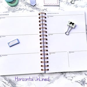 May include: A spiral-bound planner open to a weekly spread for November. The pages are white with a horizontal layout. A gold pen, a binder clip with a black bow, and sticky notes are on the marble surface. The text "Horizontal Unlined" is at the bottom.
