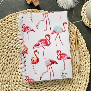 Op de afbeelding: Een spiraalgebonden notitieboek met een witte kaft voorzien van een herhalend patroon van roze flamingo's. In de rechterbenedenhoek staan de naam Lisa en het jaartal 2026 in zwarte letters. Een goudkleurige pen met kristallen ligt ernaast.