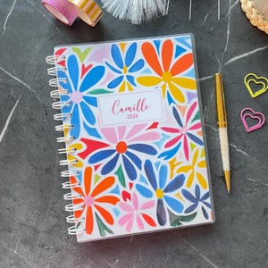 Op de afbeelding: Een spiraalgebonden agenda met een kleurrijk bloemenontwerp. De omslag heeft een levendig patroon van blauwe, oranje, gele en roze bloemen. De naam "Camille" en het jaar "2026" staan op een wit label. Een gouden pen en hartvormige paperclips zijn ook zichtbaar.