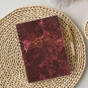 May include: A spiral-bound notebook with a dark burgundy and black marble pattern cover. The name Janine and the year 2024 are printed in gold script on the front. A gold-toned pen with a clear crystal-filled barrel is placed next to the notebook.