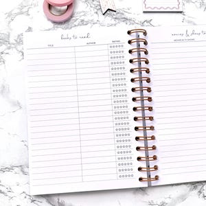 May include: Open spiral notebook with lined pages for tracking books and movies. The left page is labeled "books to read" with columns for title, author, and rating. The right page is for "movies & shows to" with a star rating system. Pink washi tape and a decorative tag are visible.