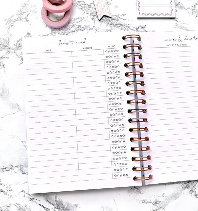 May include: Open spiral notebook with lined pages for tracking books and movies. The left page is labeled "books to read" with columns for title, author, and rating. The right page is for "movies & shows to" with a star rating system. Pink washi tape and a decorative tag are visible.