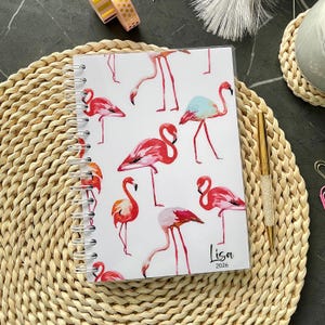 Peut inclure: Un carnet à spirales avec une couverture blanche ornée d'un motif de flamants roses et rouges. Le carnet porte le nom "Lisa" et l'année "2026". Un stylo doré avec un corps incrusté de cristaux est posé à côté.