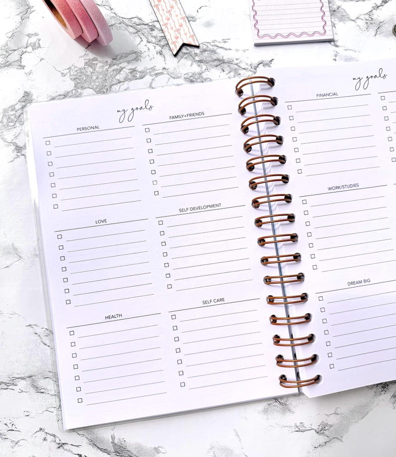 May include: Open planner with a copper spiral binding, featuring sections for goals like 'Personal,' 'Family + Friends,' and 'Love.' The pages are white with a marble-like background. The words 'my goals' are written at the top.