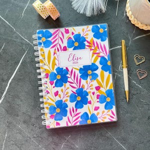 Può includere: Un taccuino a spirale con copertina bianca caratterizzato da un vivace motivo di fiori blu, foglie gialle e steli rosa. Un'etichetta rettangolare centrale riporta il testo Elise 2026 in un carattere corsivo su una superficie scura.