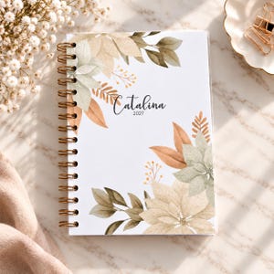 Könnte beinhalten: Ein spiralgebundenes Notizbuch mit weißem Einband und einem Aquarell-Blumenmuster in gedeckten Beige-, Salbeigrün- und sanften Orangetönen. In der Mitte des Einbands stehen der Name Catalina und das Jahr 2027 in schwarzer Schreibschrift.