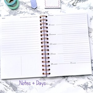May include: Open spiral-bound planner with lined pages, featuring the month of May and daily sections from Monday to Sunday. A gold pen with a crystal-like barrel rests on the side. The words "Notes + Days" are at the bottom.