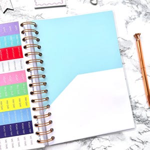 May include: Open spiral-bound planner with a light blue cover and white interior pages. The planner has gold-colored spiral binding and a matching pen with a diamond-shaped top. Colorful tabs label sections for appointments, meetings, deadlines, and more.
