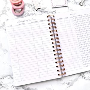 May include: Open notebook with a spiral binding and a subscriptions tracker layout. The pages are white with gray lines and headings. Pink washi tape rolls, a to-do note, and a paper clip are on the marble surface.