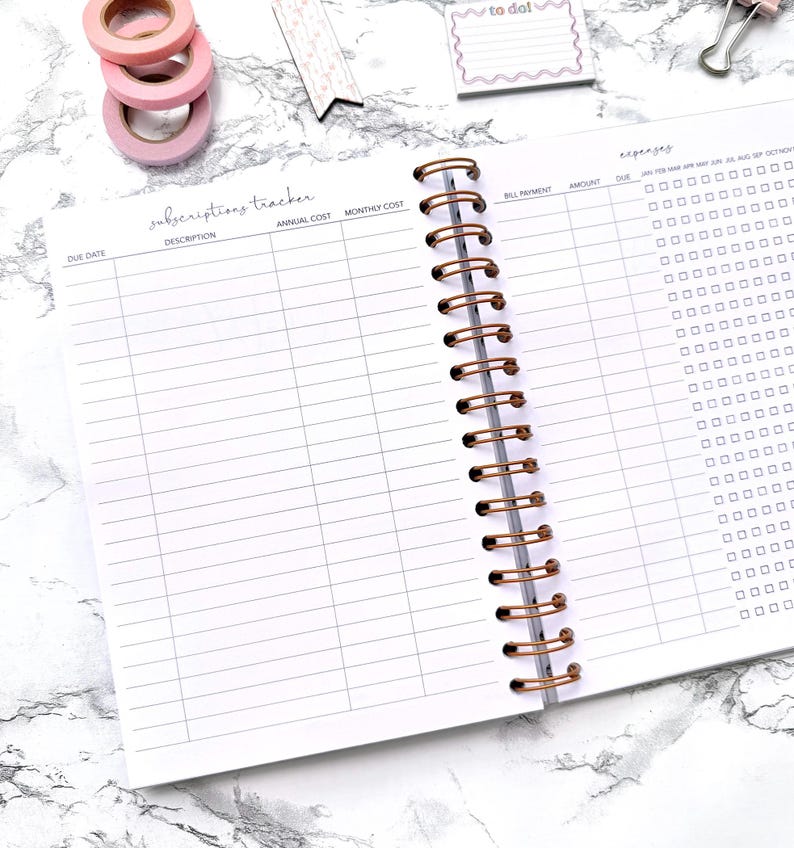 May include: Open notebook with a spiral binding and a subscriptions tracker layout. The pages are white with gray lines and headings. Pink washi tape rolls, a to-do note, and a paper clip are on the marble surface.