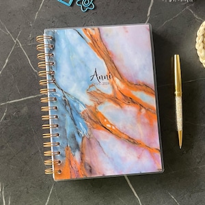 May include: A spiral-bound planner with a marble-like cover in shades of blue, orange, and white. The cover reads "Anni 2025-2026". A gold pen with a crystal-studded barrel rests beside the planner. The planner's binding is gold.
