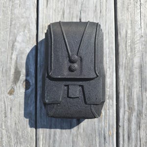 以下が含まれることがあります： フラップと2つのボタン留めが付いた、濃い灰色の長方形のケース。ケースはテクスチャ加工された表面と、前面にストラップのようなデザインが施されています。ケースは風化した木製の表面に置かれています。