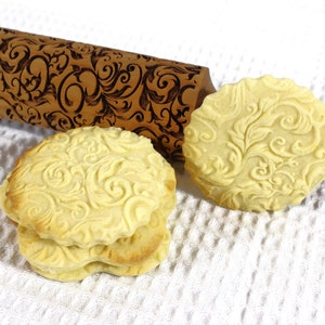 Könnte beinhalten: Ein Holz-Nudelholz mit einem dekorativen Blumenmuster ist mit zwei Keksen abgebildet, die mit dem Nudelholz gerollt wurden. Die Kekse sind hellgelb und haben ein erhabenes Blumenmuster.