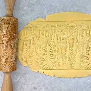 May include: A wooden rolling pin with a carved design of a forest scene is used to roll out a sheet of dough. The dough is imprinted with the design of the rolling pin, creating a detailed forest scene.