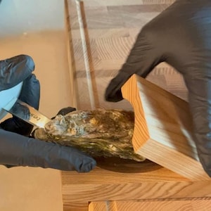 Op de afbeelding: Een persoon met zwarte handschoenen opent een oester met een oestermes. De oester ligt in een houten oesteropener.