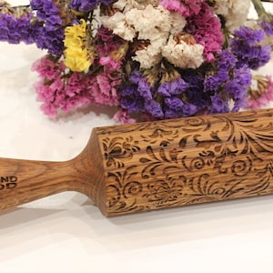 May include: A wooden rolling pin with an intricate floral design engraved into the surface. The rolling pin has a brown wooden handle with the words "DIAMOND WOOD" engraved into it.