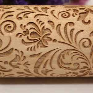 May include: A close-up of a wooden rolling pin with an intricate floral design carved into the surface. The design features swirls, leaves, and flowers.