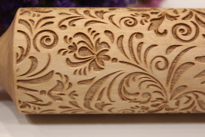 May include: A close-up of a wooden rolling pin with an intricate floral design carved into the surface. The design features swirls, leaves, and flowers.