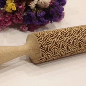 May include: A wooden rolling pin with a geometric pattern carved into the surface. The rolling pin is made of light brown wood and has a handle.