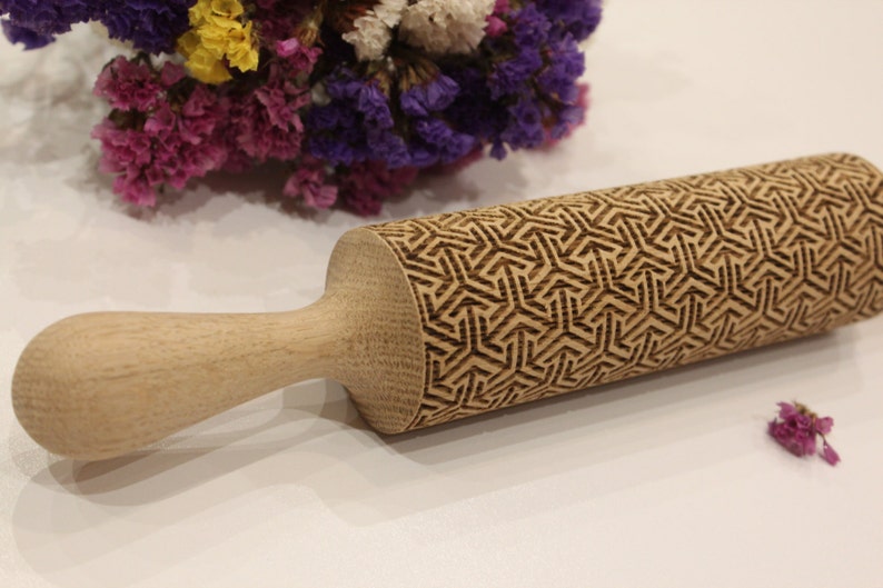 May include: A wooden rolling pin with a geometric pattern carved into the surface. The rolling pin is made of light brown wood and has a handle.