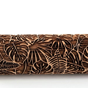 Op de afbeelding: Een houten deegroller met een gedetailleerd, gesneden tropisch bladerpatroon. Het ontwerp omvat grote monstera bladeren, varens en ander gebladerte. De deegroller is gemaakt van lichtbruin hout.