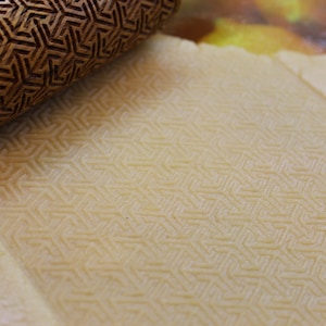 May include: A close-up of a wooden rolling pin with a geometric pattern pressed into a sheet of dough. The dough has a light beige color and is textured with the pattern from the rolling pin.