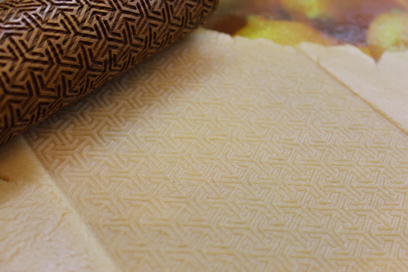 May include: A close-up of a wooden rolling pin with a geometric pattern pressed into a sheet of dough. The dough has a light beige color and is textured with the pattern from the rolling pin.