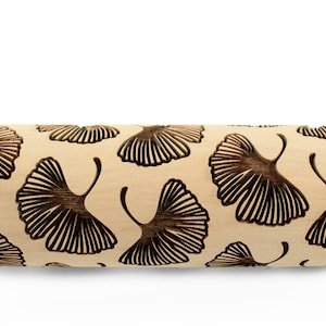 May include: A light-colored wooden rolling pin with a repeating pattern of dark brown ginkgo leaf designs. The rolling pin is cylindrical with rounded handles on each end. The ginkgo leaf pattern is carved into the wood.