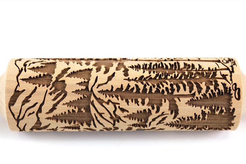 May include: A wooden rolling pin with a detailed forest scene carved into its surface. The design features trees and foliage, creating a textured pattern. The rolling pin is light brown with a natural wood grain.