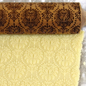 May include: A wooden rolling pin with an ornate floral pattern is rolling over a sheet of dough, creating an embossed design. The rolling pin is brown, and the dough is a light yellow color. The scene suggests baking or pastry making.