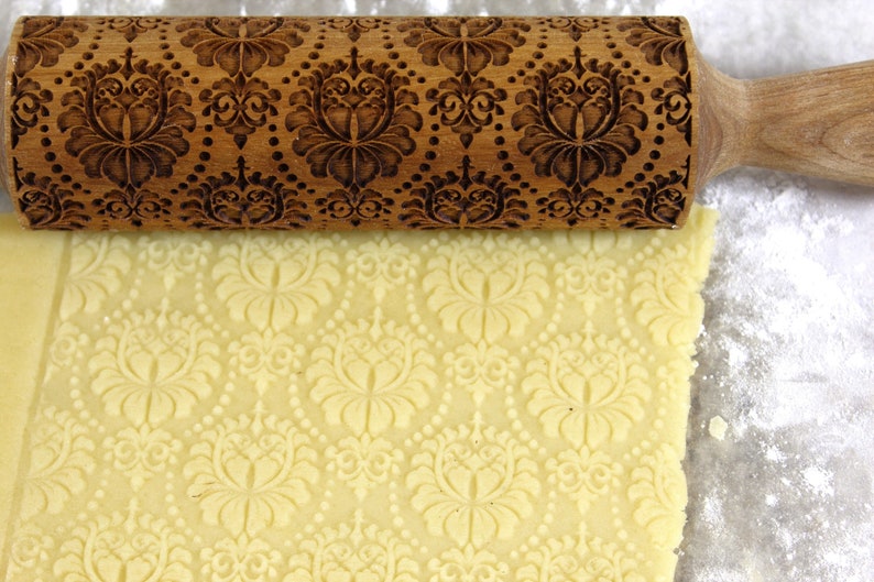 May include: A wooden rolling pin with an ornate floral pattern is rolling over a sheet of dough, creating an embossed design. The rolling pin is brown, and the dough is a light yellow color. The scene suggests baking or pastry making.