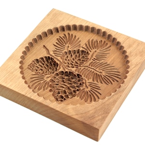 Puede incluir: Un cortador de galletas de madera con un dise&ntilde;o detallado de pi&ntilde;as y ramas. El cortador es de forma cuadrada con bordes redondeados y tiene un borde festoneado alrededor del dise&ntilde;o.