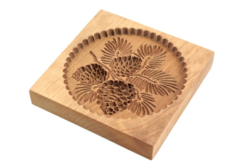 Puede incluir: Un cortador de galletas de madera con un dise&ntilde;o detallado de pi&ntilde;as y ramas. El cortador es de forma cuadrada con bordes redondeados y tiene un borde festoneado alrededor del dise&ntilde;o.