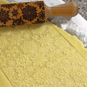 May include: A wooden rolling pin with a snowflake design is used to roll out dough for cookies. The dough is imprinted with the snowflake design.
