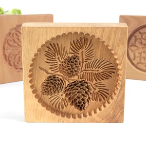 Puede incluir: Cuatro cortadores de galletas de madera, cada uno con un dise&ntilde;o diferente. El cortador del centro presenta un dise&ntilde;o de pi&ntilde;a y agujas de pino. Los otros cortadores presentan un dise&ntilde;o floral.