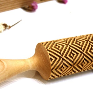 Può includere: Un mattarello di legno con un motivo geometrico inciso sulla superficie. Il motivo è composto da quadrati e triangoli ripetuti.