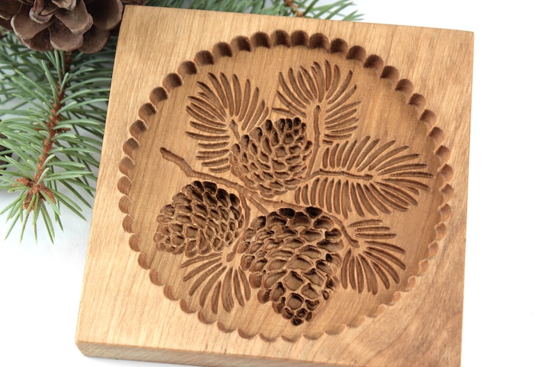 Puede incluir: Un cortador de galletas de madera con un dise&ntilde;o detallado de tres pi&ntilde;as y agujas de pino. El cortador tiene un borde festoneado.