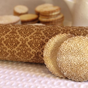 May include: A wooden rolling pin with a brown and gold damask pattern. The rolling pin is lying on a white surface with two decorative cookies with a similar damask pattern.