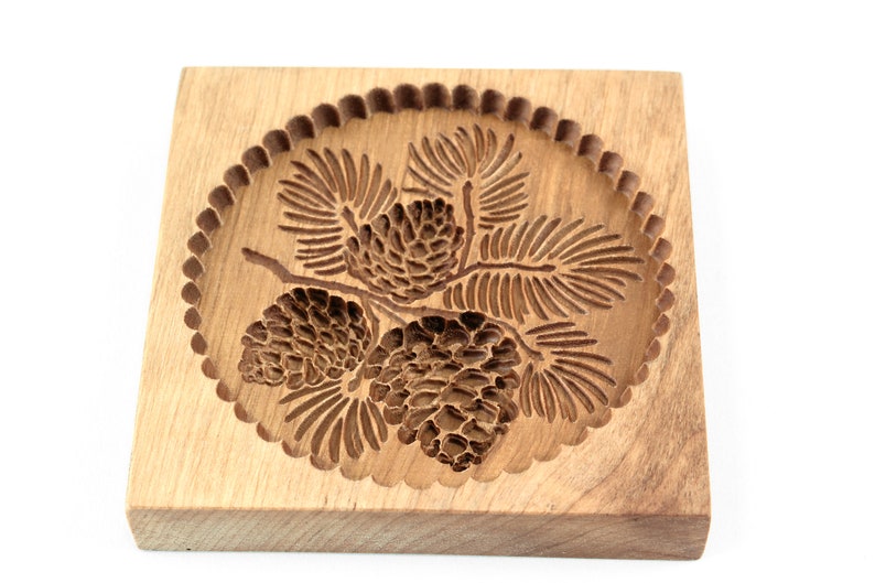 Puede incluir: Un cortador de galletas de madera con un dise&ntilde;o detallado de pi&ntilde;as y ramas. El cortador es redondo con un borde festoneado.