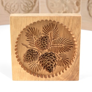 Puede incluir: Cortador de galletas de madera con un dise&ntilde;o de pi&ntilde;a. El cortador es de forma cuadrada y tiene un dise&ntilde;o circular en el centro con tres pi&ntilde;as y agujas de pino.