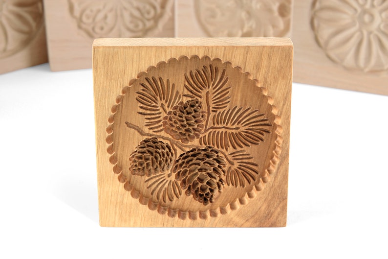 Puede incluir: Cortador de galletas de madera con un dise&ntilde;o de pi&ntilde;a. El cortador es de forma cuadrada y tiene un dise&ntilde;o circular en el centro con tres pi&ntilde;as y agujas de pino.