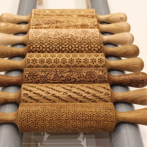 May include: A set of nine wooden rolling pins with various engraved designs. The rolling pins are arranged in a row, with the handles pointing to the right. The designs include snowflakes, floral patterns, and geometric shapes. Some of the rolling pins have personalized messages engraved on them, such as "Made with love by Rebecca".