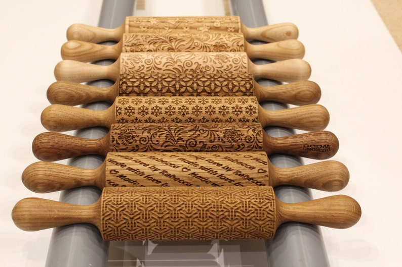 May include: A set of nine wooden rolling pins with various engraved designs. The rolling pins are arranged in a row, with the handles pointing to the right. The designs include snowflakes, floral patterns, and geometric shapes. Some of the rolling pins have personalized messages engraved on them, such as "Made with love by Rebecca".