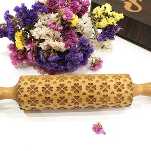 May include: A wooden rolling pin with a carved pattern of hearts and clover leaves. The rolling pin is made of light brown wood and has a natural finish.