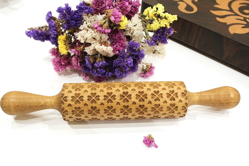 May include: A wooden rolling pin with a carved pattern of hearts and clover leaves. The rolling pin is made of light brown wood and has a natural finish.