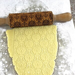 May include: A wooden rolling pin with a decorative floral pattern is shown over a sheet of dough. The dough has been rolled out and imprinted with the same floral design. The rolling pin has wooden handles.