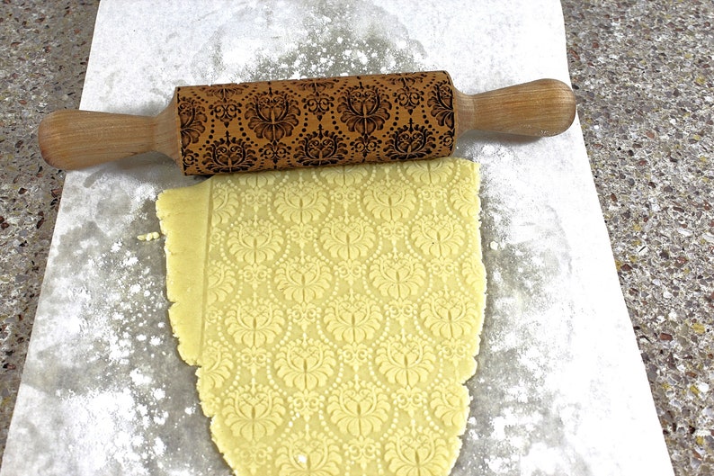 May include: A wooden rolling pin with a decorative floral pattern is shown over a sheet of dough. The dough has been rolled out and imprinted with the same floral design. The rolling pin has wooden handles.