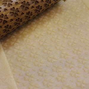 May include: A wooden rolling pin with a carved clover pattern is rolling over a sheet of dough, creating an embossed pattern of clovers on the dough.