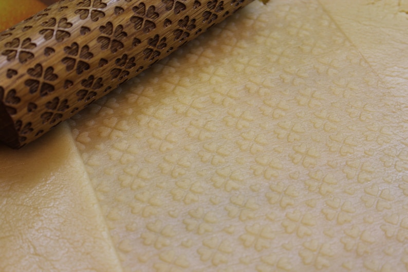 May include: A wooden rolling pin with a carved clover pattern is rolling over a sheet of dough, creating an embossed pattern of clovers on the dough.