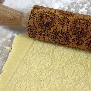 May include: A wooden rolling pin with an embossed floral pattern rests on a sheet of dough, creating a matching design. The rolling pin is light brown, and the dough is a pale yellow. Flour dusts the surface.